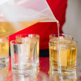 A person pouring a yellow substance, likely wax, into a small glass container, with other glasses arranged around the table as if for a candle-making workshop.