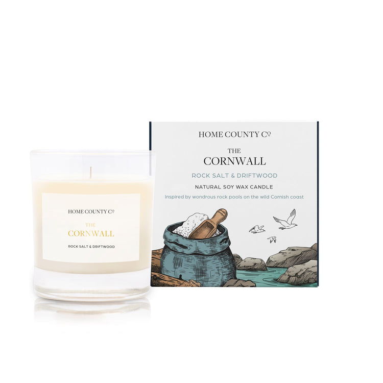 A rock salt and driftwood scented soy wax candle inspired by Cornwall is shown next to its sustainable candle box with coastal illustration