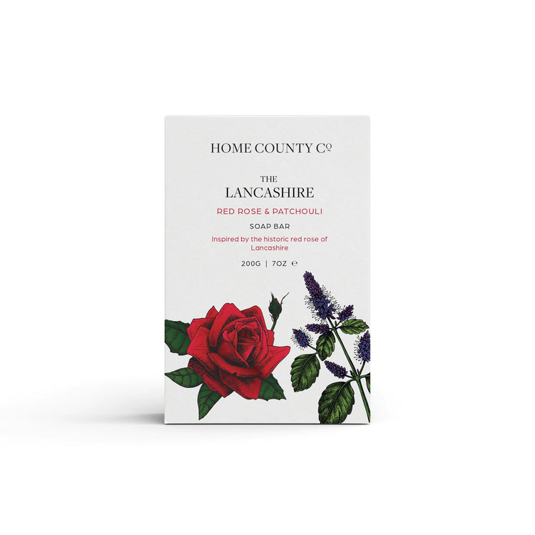 A red rose scented natural soap bar from the Home County Co. is shown in its red rose illustrated recyclable soap bar wrap