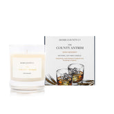 An Irish whiskey scented soy wax candle inspired by County Antrim is shown next to its sustainable candle box with whiskey illustration