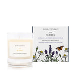 An English lavender and chamomile scented soy wax candle inspired by Surrey is shown next to its sustainable candle box with lavender illustration