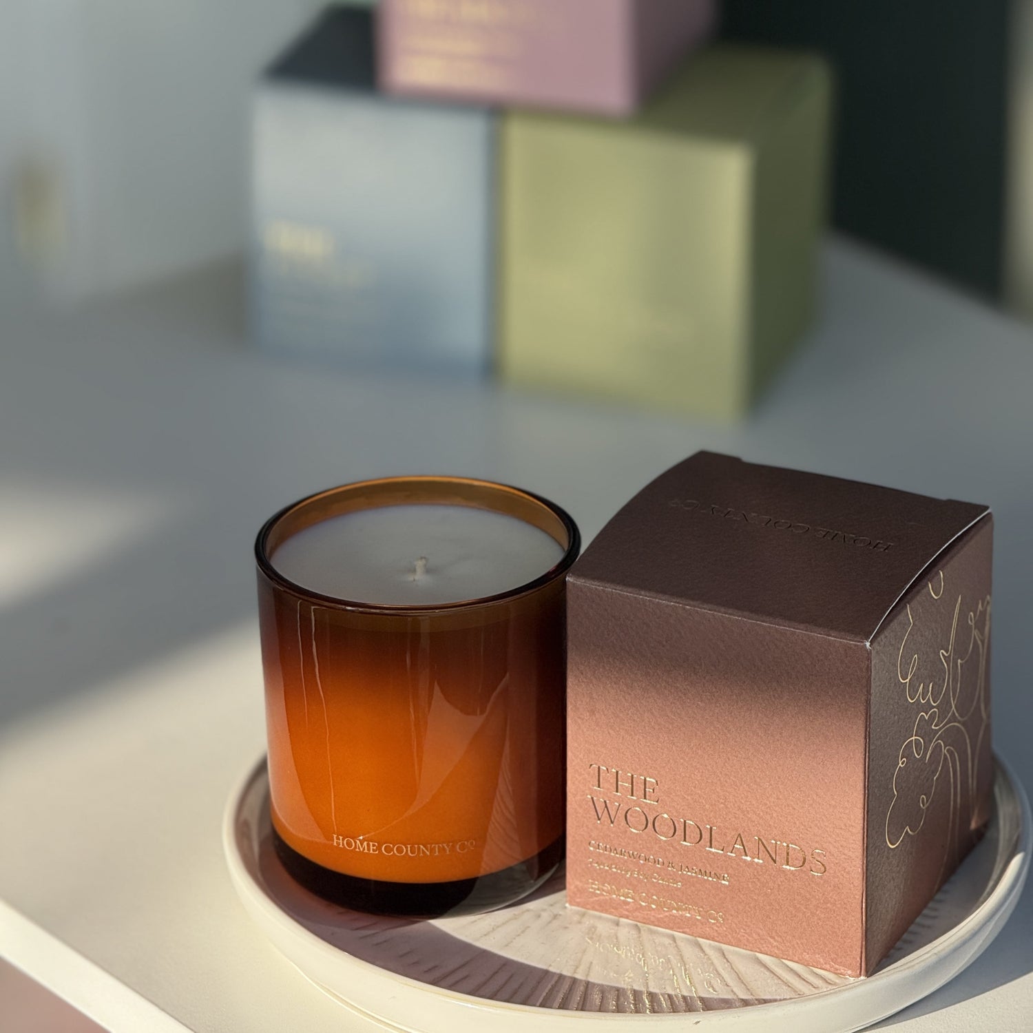 The Woodlands candle from the Home County Co. is shown in its amber candle glass vessel next to the luxury gold foiled brown box with tree emblem