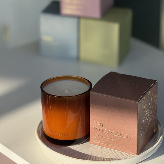 The Woodlands candle from the Home County Co. is shown in its amber candle glass vessel next to the luxury gold foiled brown box with tree emblem
