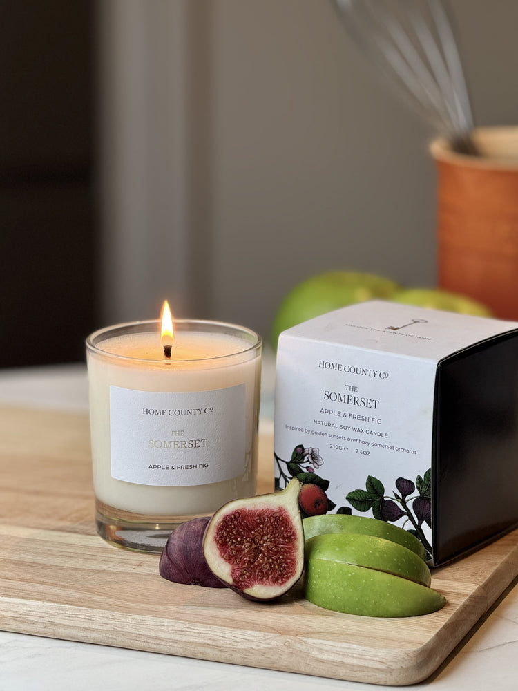 Somerset candle by Home County Co. is lit and shown next to its candle box with apple crumble in foreground