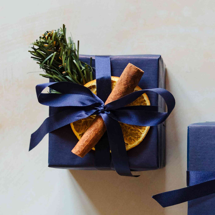 A coastal blossom scented 200g soy candle from the Home County Co. is shown with luxury Christmas Gift Wrap. The candle is wrapped in luxury navy wrapping paper secured with navy ribbon and Christmas embellishments.