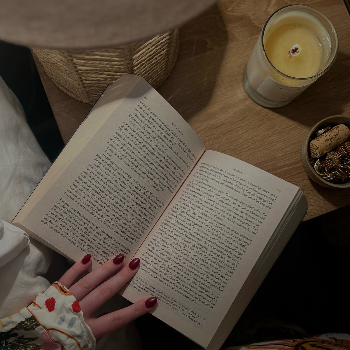 An open book with printed text is held by a person wearing a patterned pajama sleeve with red nail polish, set on a wooden table next to a lit candle and a lamp.