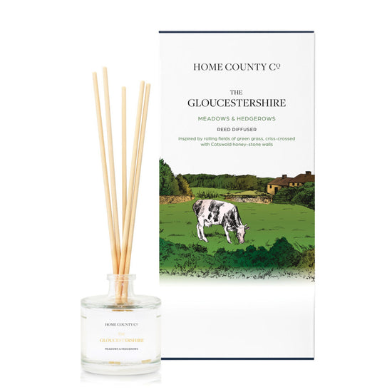 A freshly cut grass scented reed diffuser from Home County Co. The vegan friendly reed diffuser is shown next to the eco friendly reed diffuser box packaging with Cotswold hills illustration.