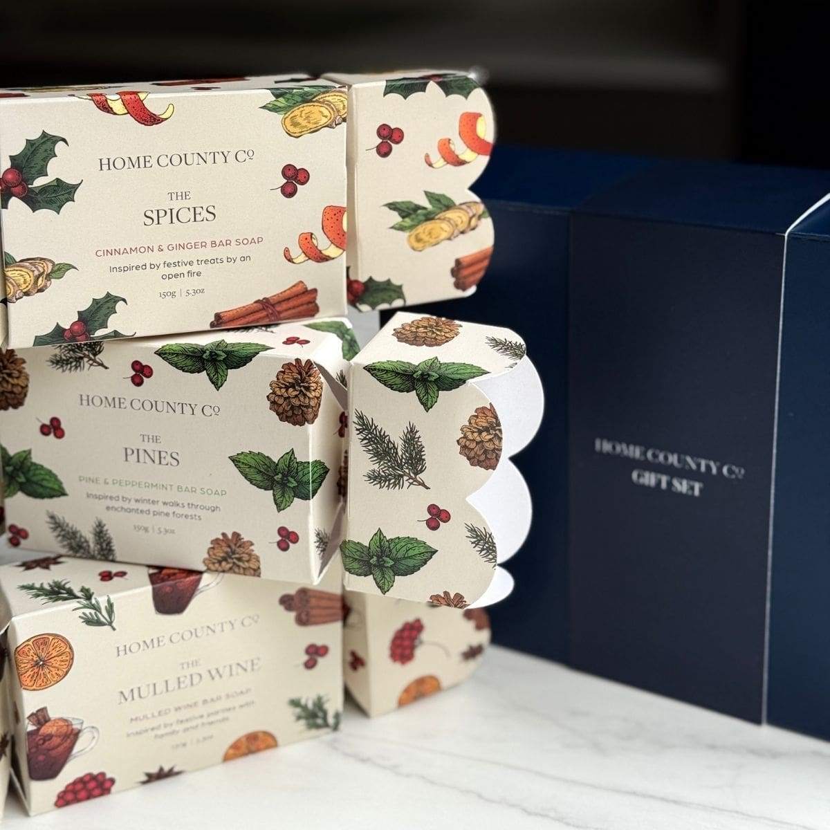 Home County Co. Christmas Soap Bar Crackers are shown stacked on top of each other in front of the luxury navy gift box