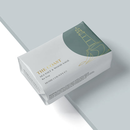 A coastal sea salt and wood sage soap bar from the Home County Co. is shown wrapped in a luxury soap wrap with gold foil lighthouse illustration
