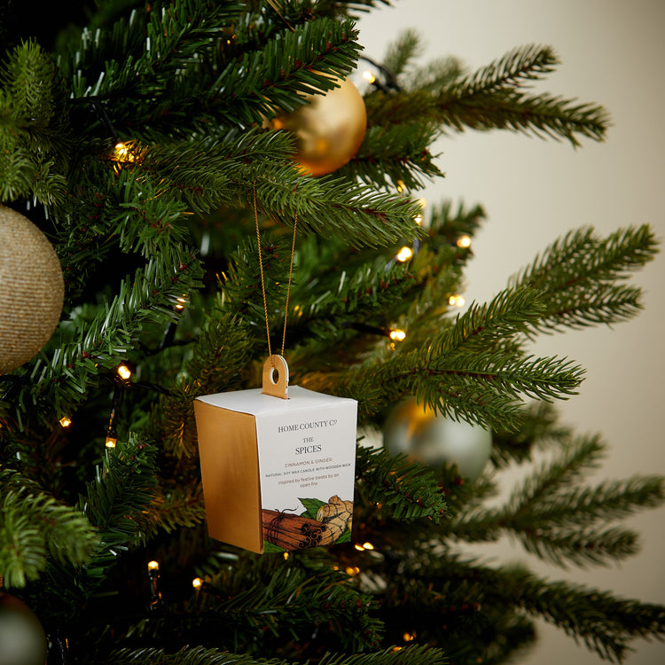 A cinnamon and ginger votive Christmas candle from the Home County Co. is shown in eco-friendly packaging hanging on the Christmas tree