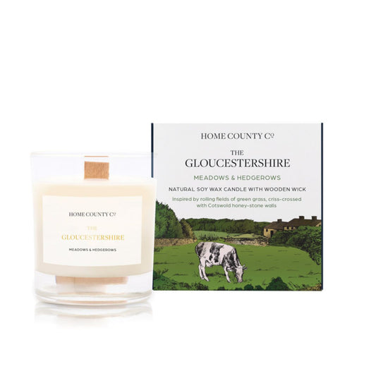 A freshly cut grass scented candle from Home County Co. The wooden wick soy candle is shown next to the eco friendly candle box packaging which displays an illustration of Cotswold hills.