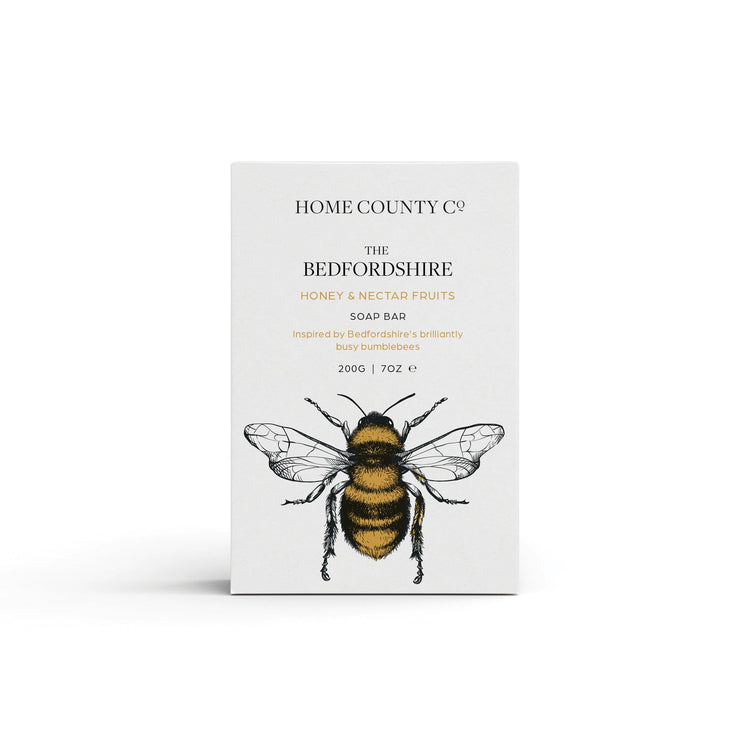 A honey scented natural soap bar from the Home County Co. is shown in its honey bee illustrated recyclable soap bar wrap