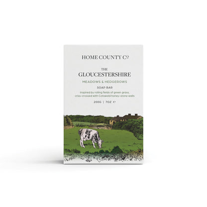 A fresh cut grass scented natural soap bar from the Home County Co. is shown in its meadows illustrated recyclable soap bar wrap