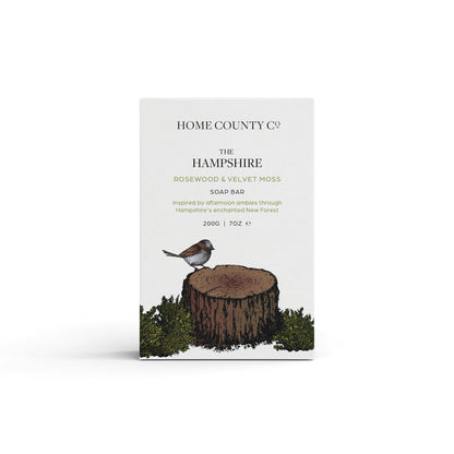 A rosewood and velvet moss woody scented natural soap bar from the Home County Co. is shown in its woodland illustrated recyclable soap bar wrap