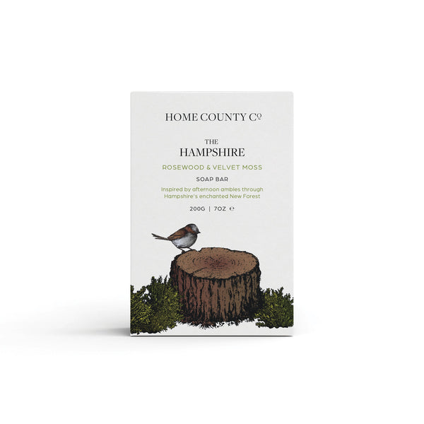 A rosewood and velvet moss woody scented natural soap bar from the Home County Co. is shown in its woodland illustrated recyclable soap bar wrap