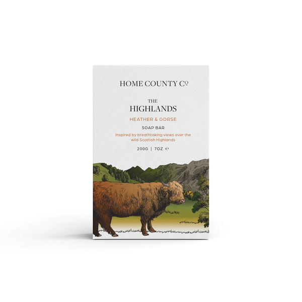 A heather and gorse scented natural soap bar from the Home County Co. is shown in its Highland Cow illustrated recyclable soap bar wrap