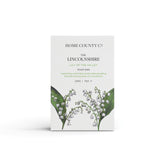 A lily of the valley scented natural soap bar from the Home County Co. is shown in its lily of the valley illustrated recyclable soap bar wrap