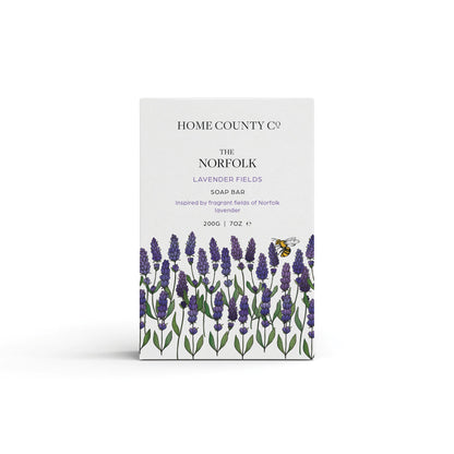 A Norfolk lavender scented natural soap bar from the Home County Co. is shown in its lavender illustrated recyclable soap bar wrap
