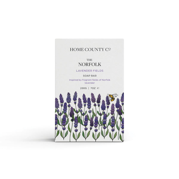 A Norfolk lavender scented natural soap bar from the Home County Co. is shown in its lavender illustrated recyclable soap bar wrap