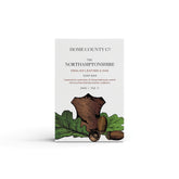 A leather and oak scented natural soap bar from the Home County Co. is shown in its leather illustrated recyclable soap bar wrap