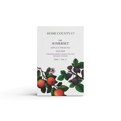 An apple and fresh fig scented natural soap bar from the Home County Co. is shown in its apple illustrated recyclable soap bar wrap