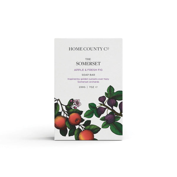 An apple and fresh fig scented natural soap bar from the Home County Co. is shown in its apple illustrated recyclable soap bar wrap