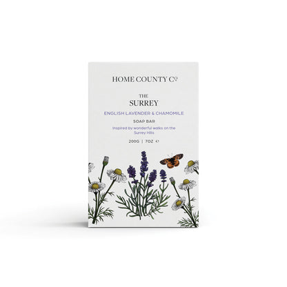 An english lavender and chamomile scented natural soap bar from the Home County Co. is shown in its lavender illustrated recyclable soap bar wrap