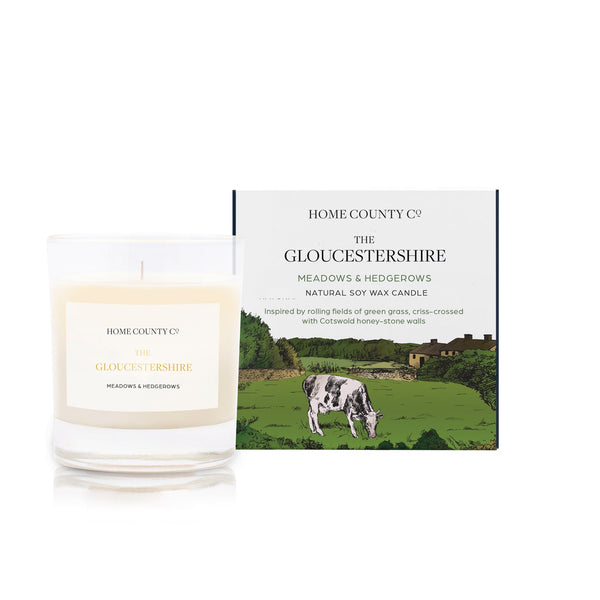 A meadows and hedgerows scented soy wax candle inspired by Gloucestershire is shown next to its sustainable candle box with Cotswold hills illustration