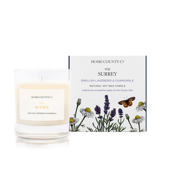An English lavender and chamomile scented soy wax candle inspired by Surrey is shown next to its sustainable candle box with lavender illustration