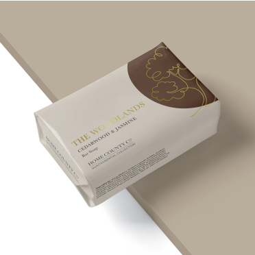 A woody cedarwood and jasmine scented soap bar from the Home County Co. is shown wrapped in a luxury soap wrap with gold foil tree illustration