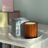 The Coast candle from the Home County Co. is shown in its amber candle glass vessel next to the luxury gold foiled blue box with lighthouse emblem
