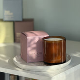 The Meadows candle from the Home County Co. is shown in its amber candle glass vessel next to the luxury gold foiled pink box with peony emblem