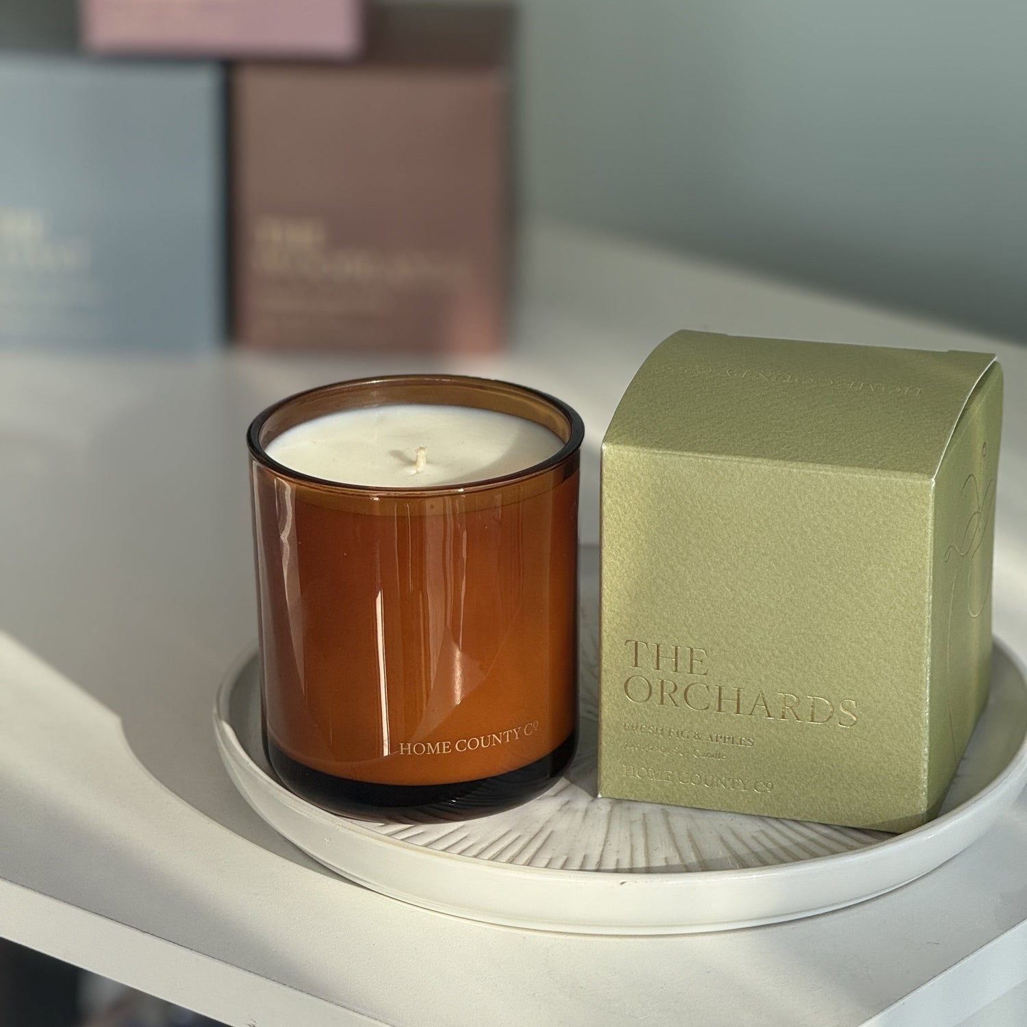 The Orchards candle from the Home County Co. is shown in its amber candle glass vessel next to the luxury gold foiled green box with apple emblem