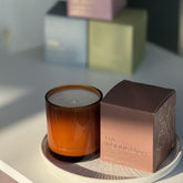 The Woodlands candle from the Home County Co. is shown in its amber candle glass vessel next to the luxury gold foiled brown box with tree emblem