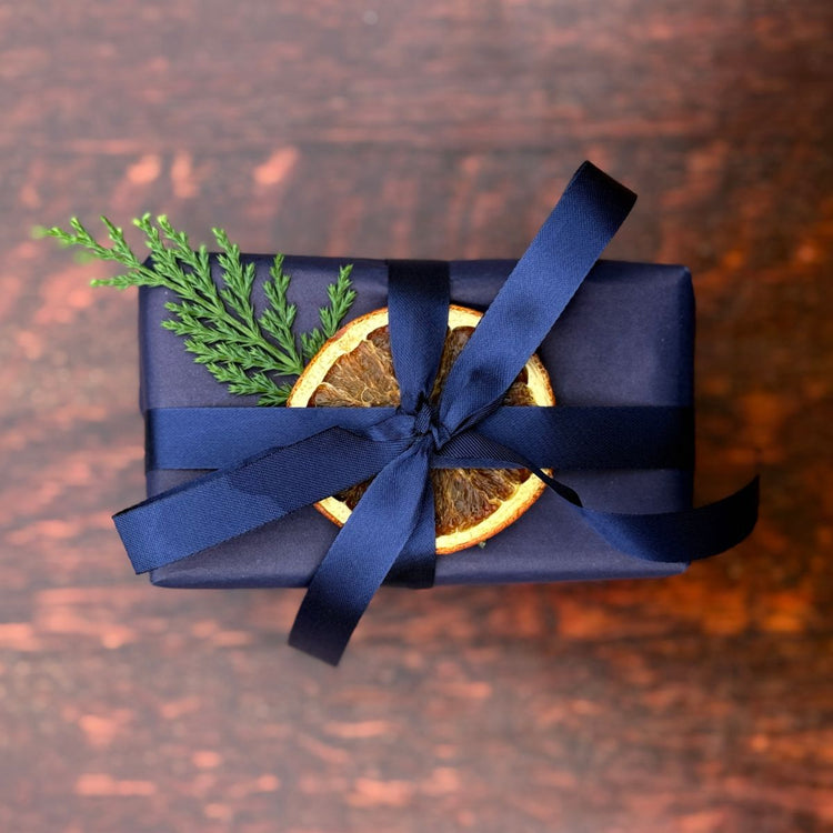 A Home County Co. soap bar is shown with Christmas gift wrap - navy wrapping paper with navy ribbon and a dried orange slice and Christmas foliage