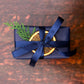A Home County Co. soap bar is shown with Christmas gift wrap - navy wrapping paper with navy ribbon and a dried orange slice and Christmas foliage