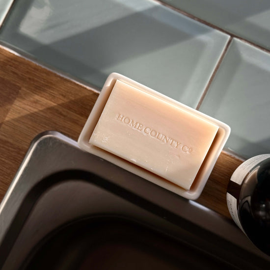 A Home County Co. county soap bar is shown unwrapped, sitting in its white ceramic soap dish from Sting in the Tail next to a tiled kitchen sink.