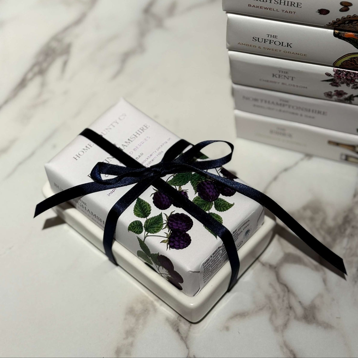A Home County Co. county soap bar is shown wrapped, sitting in its white ceramic soap dish from Sting in the Tail and held together with luxury navy ribbon for the ultimate soap gift.