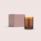 The Meadows candle from the Home County Co. is shown in its amber candle glass vessel next to the luxury gold foiled pink box with peony emblem