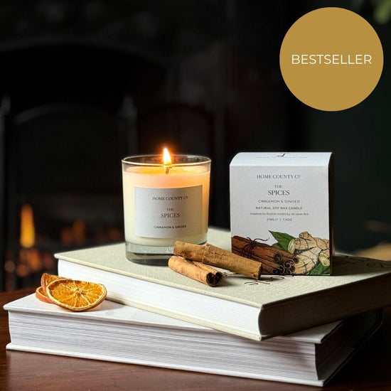 The cinnamon and ginger scented Christmas candle from the Home County Co. is shown alight next to its candle box with cinnamon sticks and orange slices in the foreground and an open fire in the background