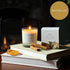 The cinnamon and ginger scented Christmas candle from the Home County Co. is shown alight next to its candle box with cinnamon sticks and orange slices in the foreground and an open fire in the background