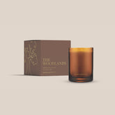 The Woodlands candle from the Home County Co. is shown in its amber candle glass vessel next to the luxury gold foiled brown box with tree emblem