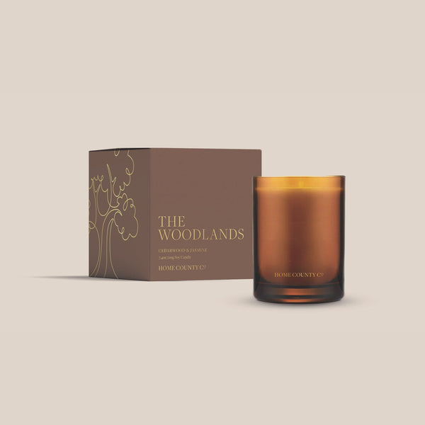 The Woodlands candle from the Home County Co. is shown in its amber candle glass vessel next to the luxury gold foiled brown box with tree emblem