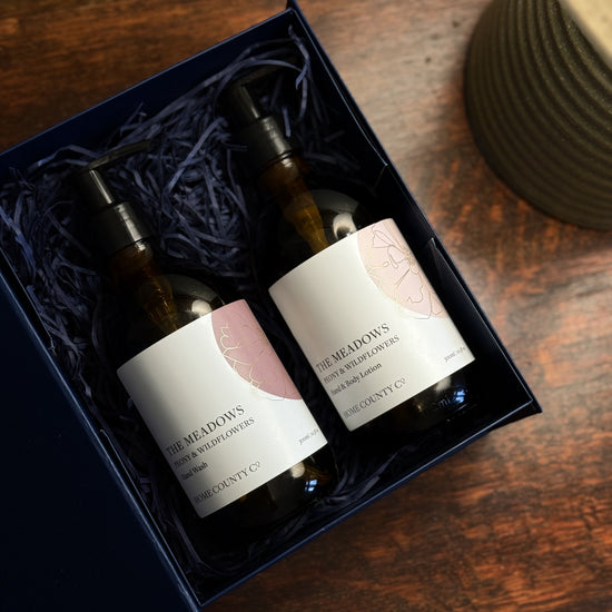 The Meadows Peony and Wildflowers liquid hand soap and lotion gift set is shown with the amber bottles in an open gift set box on a table.