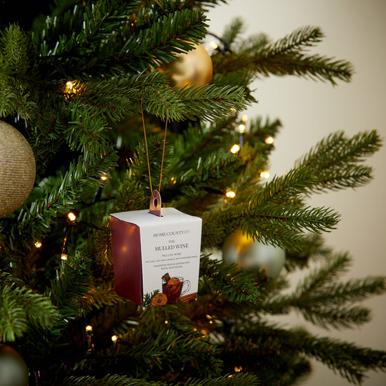 A Mulled Wine votive Christmas candle from the Home County Co. is shown in eco-friendly packaging hanging on the Christmas tree