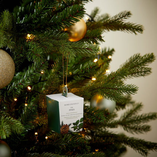 A Pine scented votive Christmas candle from the Home County Co. is shown in eco-friendly packaging hanging on the Christmas tree