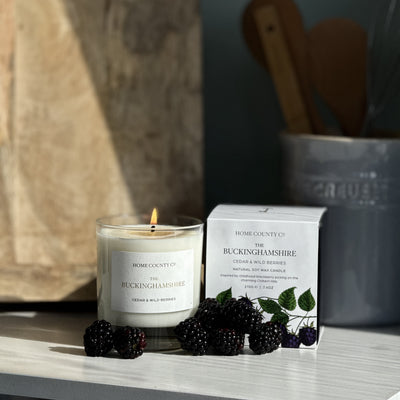 Candle and packaging labelled 'Buckinghamshire' on a surface with a dark background and blackberries in the foreground