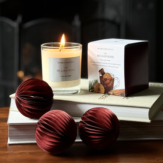 The Mulled Wine scented candle from the Home County Co. is shown alight next to its candle box with Christmas baubles and books in the foreground 