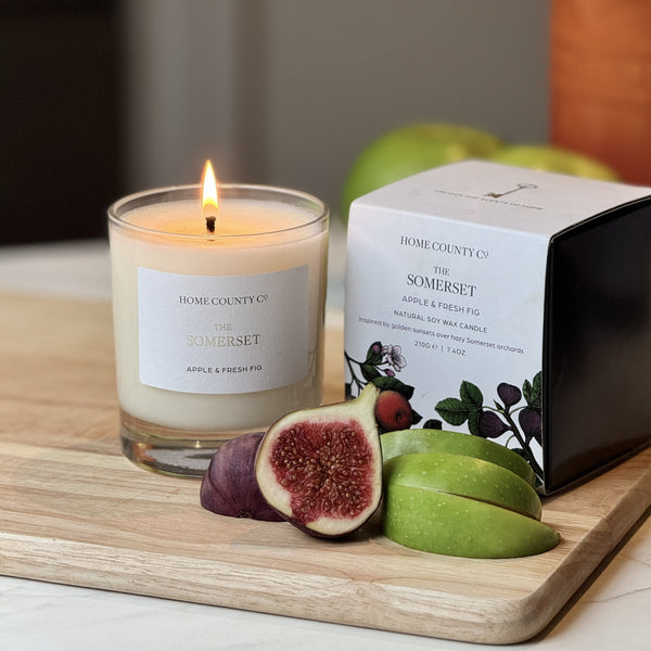 The Somerset Apple and Fresh Fig candle from Home County Co is shown alight next to its candle box with fresh apples and figs in the foreground