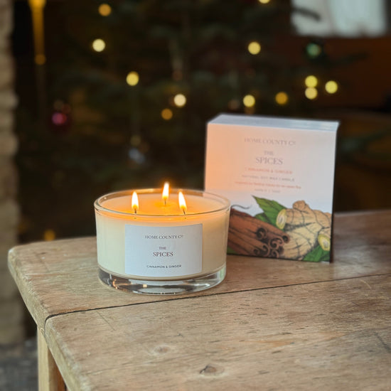 A lit scented candle in a clear glass jar sits on a rustic wooden table next to its product box. The background is a warm, softly blurred indoor setting with a glowing Christmas tree and warm lights.
